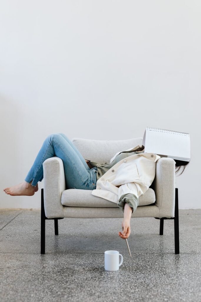 Tired woman in casual attire falls asleep on sofa with a notebook on face and pen in hand.